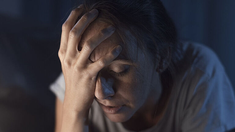Eine Frau sitzt mit geschlossenen Augen in der Dunkelheit und hält sich vor Müdigkeit den Kopf.