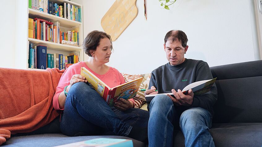 Eine Frau und ein Mann sitzen auf einem Sofa, sie hält ein Buch in den Händen, er ein Schreibheft.