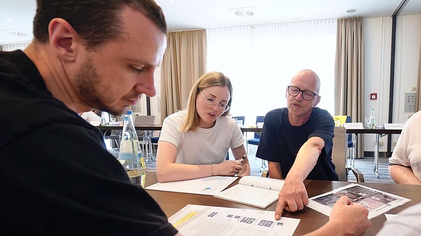 Vier Personen sitzen gemeinsam an einem Tisch. Sie betrachten Unterlagen und Fotos und diskutieren. Ein Mann deutet mit dem Finger auf ein Blatt Papier, eine Frau neben ihm schaut interessiert darauf.