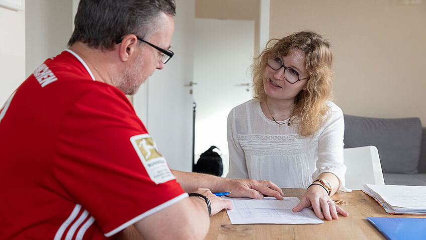 Steven Wedhorn, links im roten Trikot und Reha-Beraterin Sarah Baumgärtner, rechts von ihm, sitzen am Tisch und unterhalten sich. 