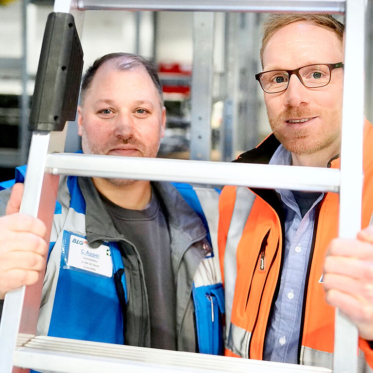 Schichtleiter Christian Appel und Thomas Menke, Leiter Operations Kommissionierung, schauen durch eine Leiter.