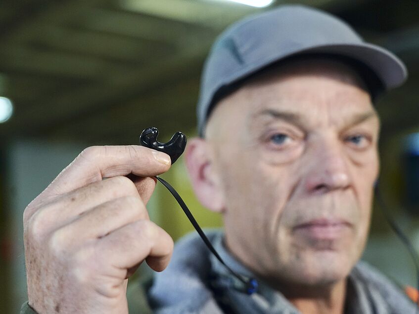 Hafenarbeiter mit Sicherheitskäppi hält maßgefertigten Gehörschutz in die Kamera.