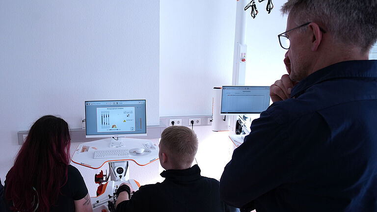 Handtherapie am Spezialgerät: Von hinten sind Ergotherapeutin Iris Stenzel, Lars Eberhard (beide sitzend) und Reha-Berater Hartmut Wahlig (stehend rechts) zu sehen. Alle schauen auf den Bildschirm des Therapie-Computers , an dem Lars seine Hände trainiert. 