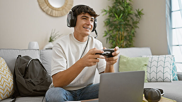 Ein junger Mann mit kurzen dunklen Haaren sitzt auf einem grauen Sofa, trägt ein Headset mit Mikrofon und hält einen Game-Controller in den Händen. Er lacht und wirkt konzentriert. Vor ihm steht ein Laptop, daneben liegt ein weiteres Headset. Im Hintergrund sind bunte Kissen, eine Pflanze und ein rundes Dekoelement an der Wand zu sehen.