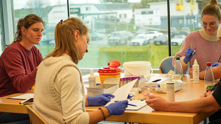 Blick in den Untersuchungsraum. Drei Ärztinnen sitzen um einen Tisch, auf dem das Equipment für die medizinischen Untersuchungen steht und sortieren und sichten das Material.