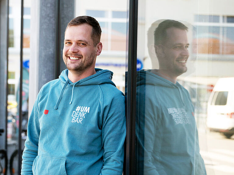  Ein Mann steht an eine Schaufensterscheibe gelehnt. In der Scheibe spiegelt sich sein Oberkörper, der ein blaues Sweatshirt trägt. Er hat die Hände in den Hosentaschen und lächelt freundlich in die Kamera.