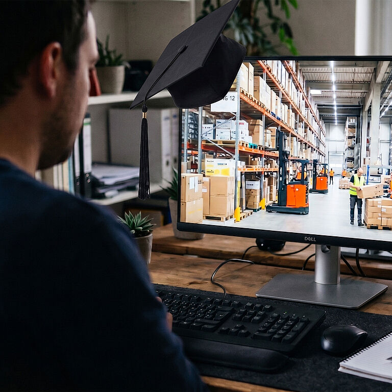 Ein Mann sitzt am Schreibtisch vor einem Monitor, darauf zu sehen: eine Lagerhalle. An dem Monitor hängt ein schwarzer Absolventenhut.
