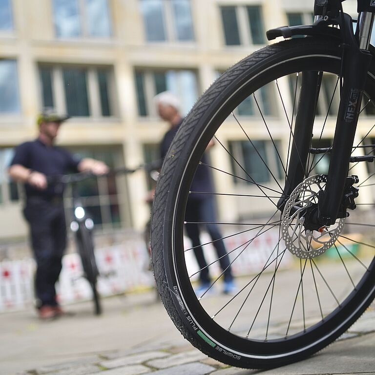 Auf einem Platz steht das Vorderrad eines E-Bikes im Vordergrund. Im Hintergrund stehen unscharf zwei Männer, der linke hält ein Fahrrad fest. Dahinter sieht man - ebenfalls unscharf - die Umrisse eines Gebäudes. 
