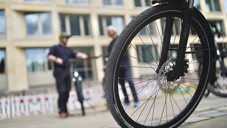 Auf einem Platz steht das Vorderrad eines E-Bikes im Vordergrund. Im Hintergrund stehen unscharf zwei Männer, der linke hält ein Fahrrad fest. Dahinter sieht man - ebenfalls unscharf - die Umrisse eines Gebäudes.
