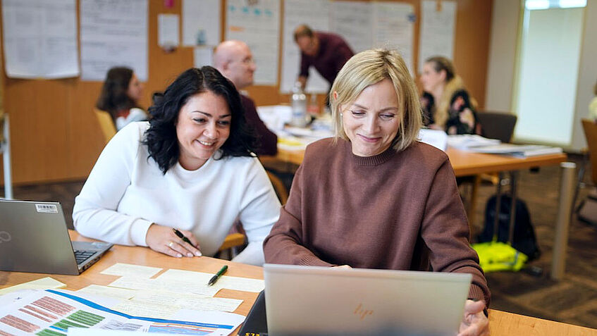 Zwei Frauen sitzen in einem Seminarraum am Tisch und schauen auf einen Laptop.