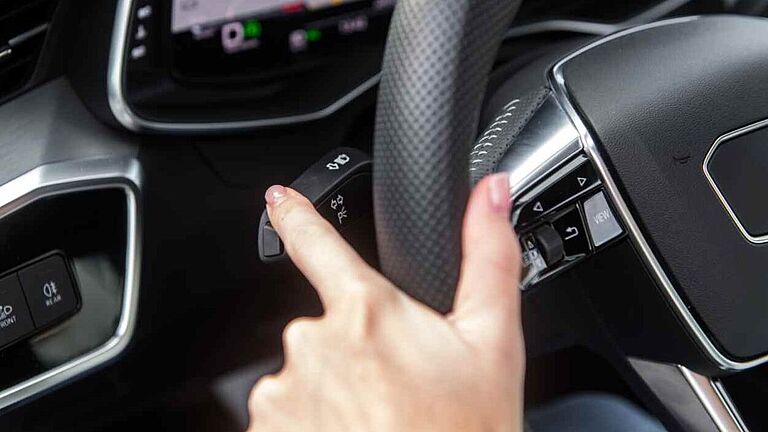 Hand am Lenkrad eines Autos betätigt den Blinkerhebel, Armaturenbrett und Navigationsanzeige im Hintergrund.