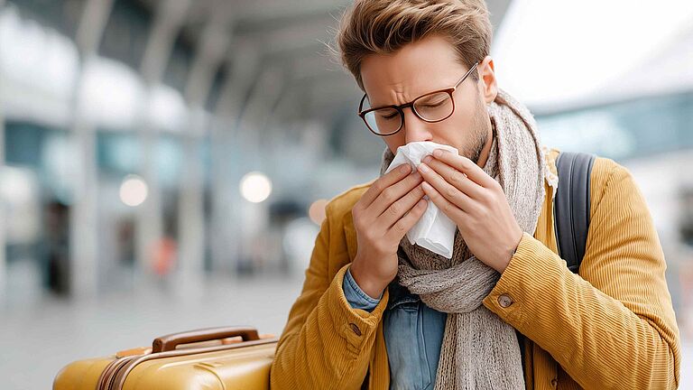Mann mit Koffer am Bahnhof niest in ein Taschentuch und wirkt erkältet.