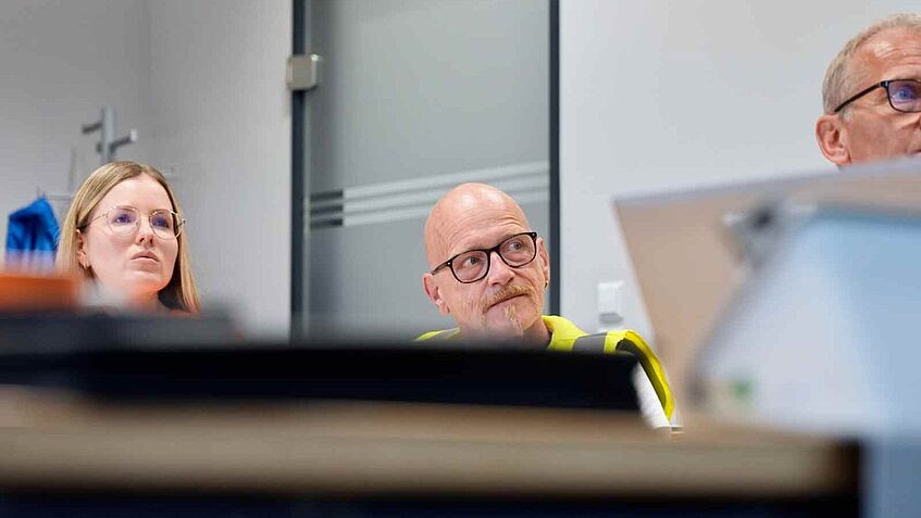 Zwei Personen sitzen in einem Besprechungsraum an einem Tisch und verfolgen eine Präsentation oder Diskussion. Im Vordergrund sind technische Geräte unscharf zu sehen.