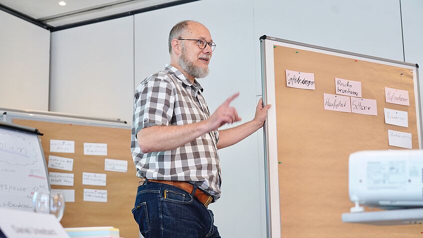 Ein Mann mit Brille und kariertem Hemd steht vor einem Flipchart und erklärt etwas. Auf dem Flipchart sind Karten mit Begriffen wie „Gefährdungen“, „Risikobeurteilung“, „Häufigkeit“ und „Schwere“ befestigt.