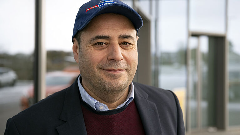 Der regionale Vertriebsleiter Ferhat Rasgele in geschäftlicher Kleidung mit Sakko, Hemd und Pullover trägt eine blaue Baseballkappe mit einem Payback-Logo. Er lächelt dezent und steht vor einer Netto-Filiale mit großen Glasfassaden. Darin spiegeln sich im Hintergrund verschwommen parkende Autos.