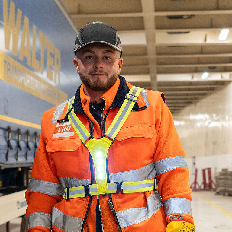 Jastin Dos Santos Costa steht in Arbeitsmontur im hell erleuchteten Schiffsdeck. Über seiner Warnweste trägt er den Warnschutzgut mit LED-Leuchten – seine Idee für mehr Arbeitssicherheit auf dem Schiff. 