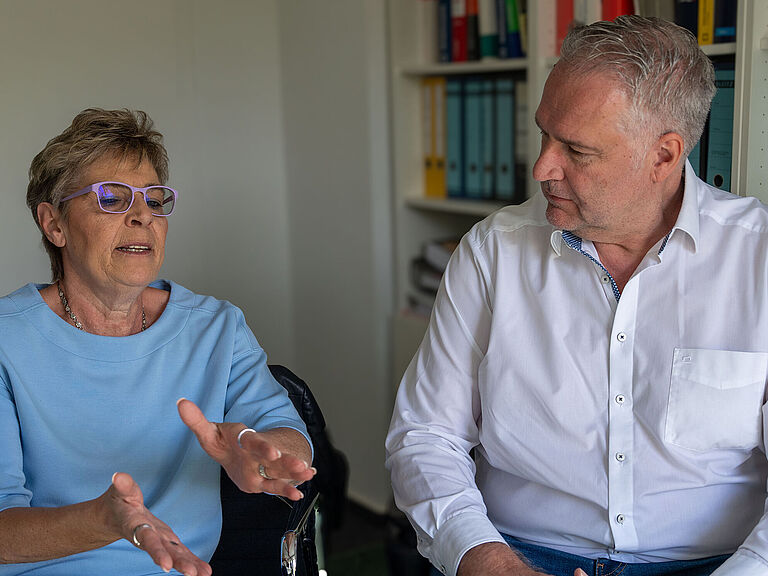 Ute Grötzinger spricht gestikulierend mit BGHW-Reha-Berater Sven Letzgus. Sie sitzen nebeneinander vor einem Regal mit Akten.