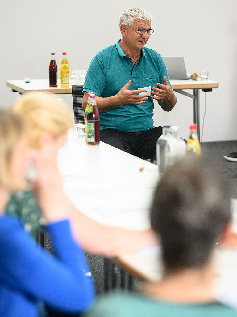 Peter Richarz vom Deutschen Rollstuhl-Sportverband (DRS) sitzt auf einem Stuhl und spricht vor Menschen, die mit dem Rücken zur Kamera zu sehen sind. Er spricht gestikulierend und wirkt konzentriert. Er trägt ein grünes T-Shirt. 