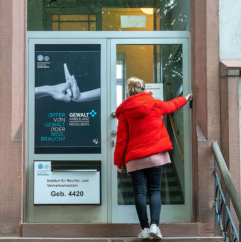 Eine Frau in leuchtend roter Jacke steht auf der Treppe vor dem Institut für Rechts- und Verkehrsmedizin in Heidelberg. Links neben der Tür hängt ein Plakat, das auf die Gewaltambulanz hinweist.