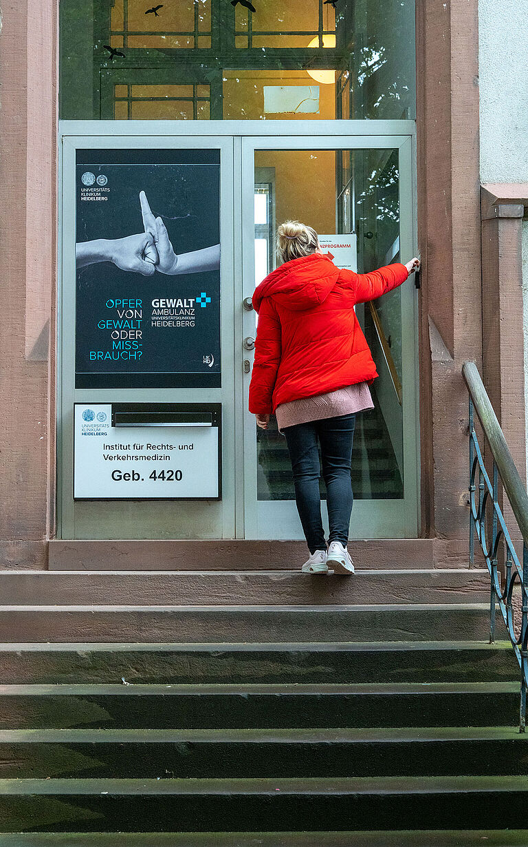 Eine Frau in leuchtend roter Jacke steht auf der Treppe vor dem Institut für Rechts- und Verkehrsmedizin in Heidelberg. Links neben der Tür hängt ein Plakat, das auf die Gewaltambulanz hinweist.