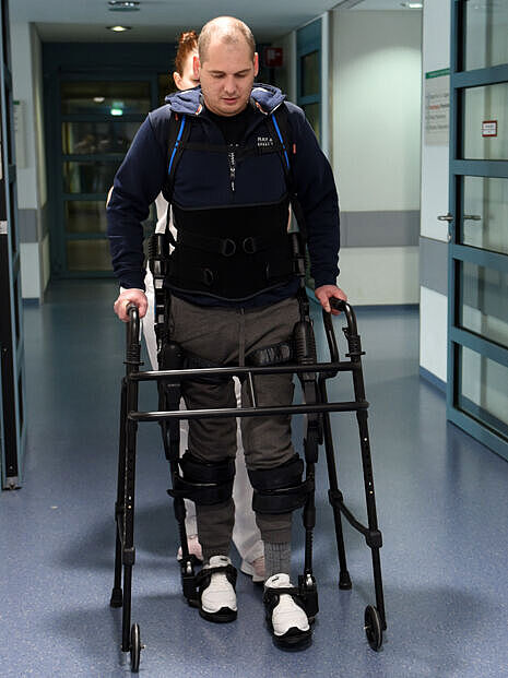 Ein Mann geht mit einem Exoskelett durch einen Krankenhausflur. Hinter ihm geht eine Therapeutin.