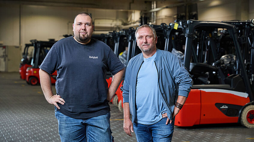 Zwei Männer stehen nebeneinander in einer Werkhalle mit roten Gabelstaplern und blicken in die Kamera.
