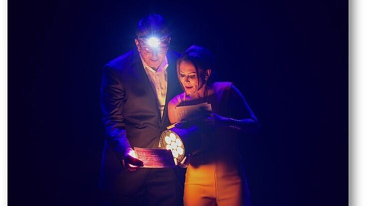 Das Moderatorenteam Tina Babbel und Karl-Josef Thielen ist kaum zu erkennen im Dunkeln. Er trägt eine Kopflampe, sie hält einen Scheinwerfer in der Hand auf ein Blatt Papier, von dem sie gemeinsam ablesen.