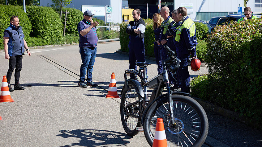 Mehrere Mitarbeitende hören zwei Trainern zu, die über das Bremsverhalten von E-Bikes aufklären. Im Vordergrund steht ein E-Bike, drei Pylone stehen auf dem Boden.