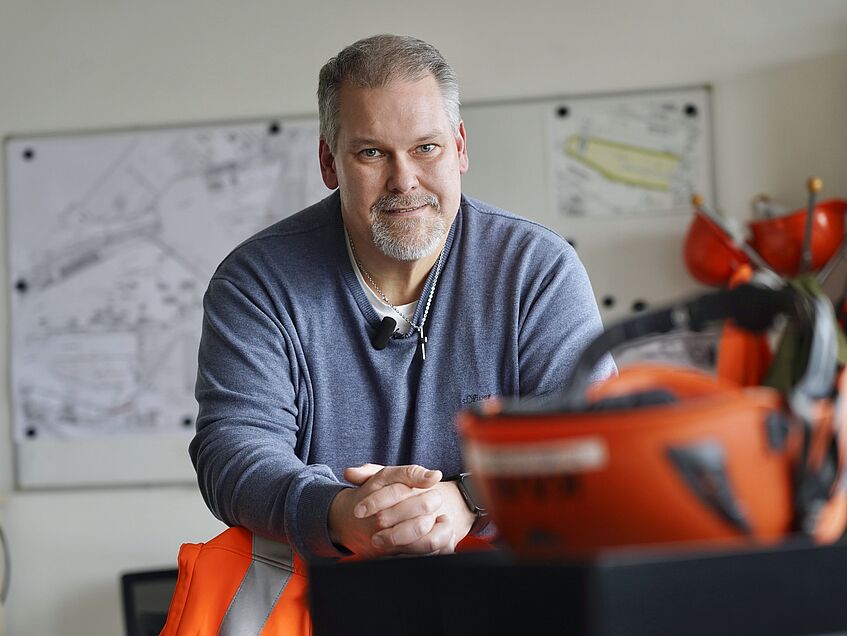 Mann sitzt im Büro vor Bauplänen und blickt in die Kamera.