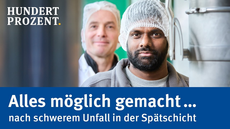 Zwei Männer stehen hintereinander und schauen zwischen zwei Metallrohren durch in die Kamera. Beide tragen weiße Hauben. Der Mann vorne hat schwarze Haare und einen Vollbart. 