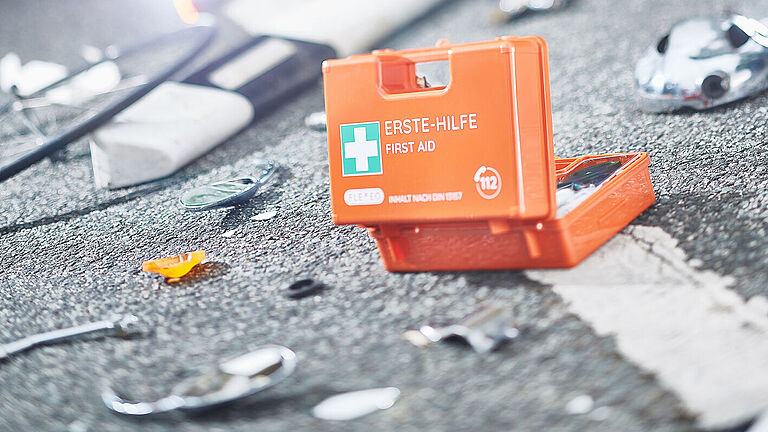 Ein leuchtend Orange-farbener Erste-Hilfe-Kasten liegt geöffnet auf dem Asphalt. Um ihn herum liegen Scherben, Splitter und ein Leitpfosten.. 