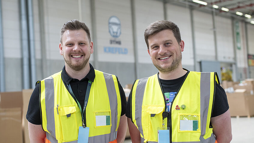 André Wilhelmi (links) und Niklas Schrage stehen mit Schutzwesten in einer Produktionshalle vom Amazon-Sortierzentrum in Krefeld und lächeln in die Kamera.