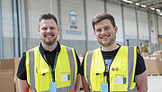 André Wilhelmi (links) und Niklas Schrage stehen mit Schutzwesten in einer Produktionshalle vom Amazon-Sortierzentrum in Krefeld und lächeln in die Kamera.