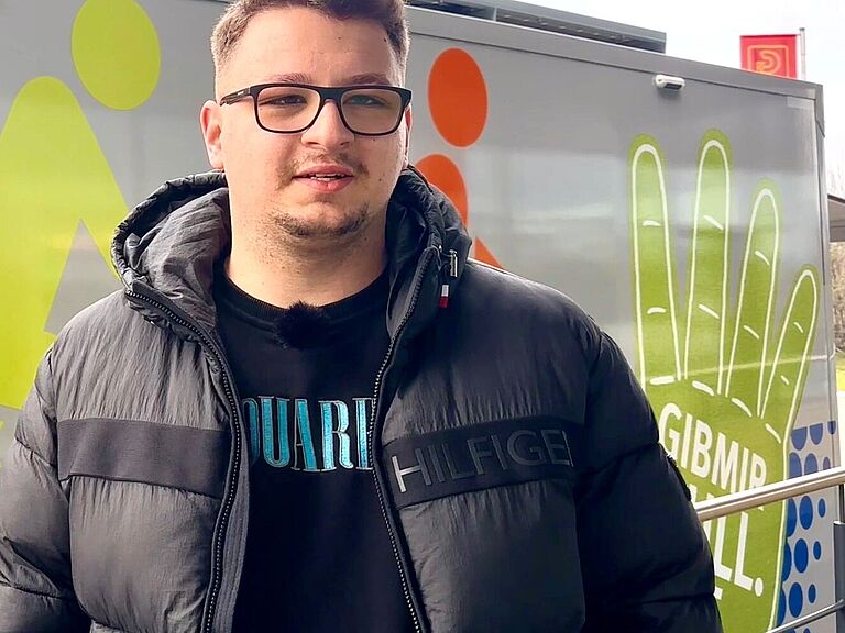 Ein junger Mann mit einer schwarzen Jacke, Brillenträger, schaut in die Kamera und lächelt. 