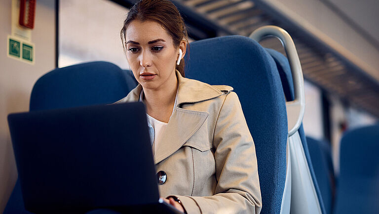 Eine junge Frau sitzt mit Laptop auf dem Schoß in der Bahn und pendelt zur Arbeit.