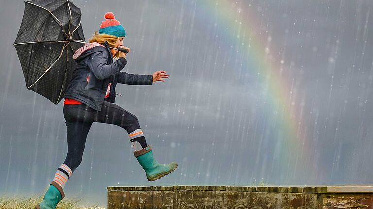 Frau mit Regenschirm geht beschwingt durch den Regen, Regenbogen am Horizont