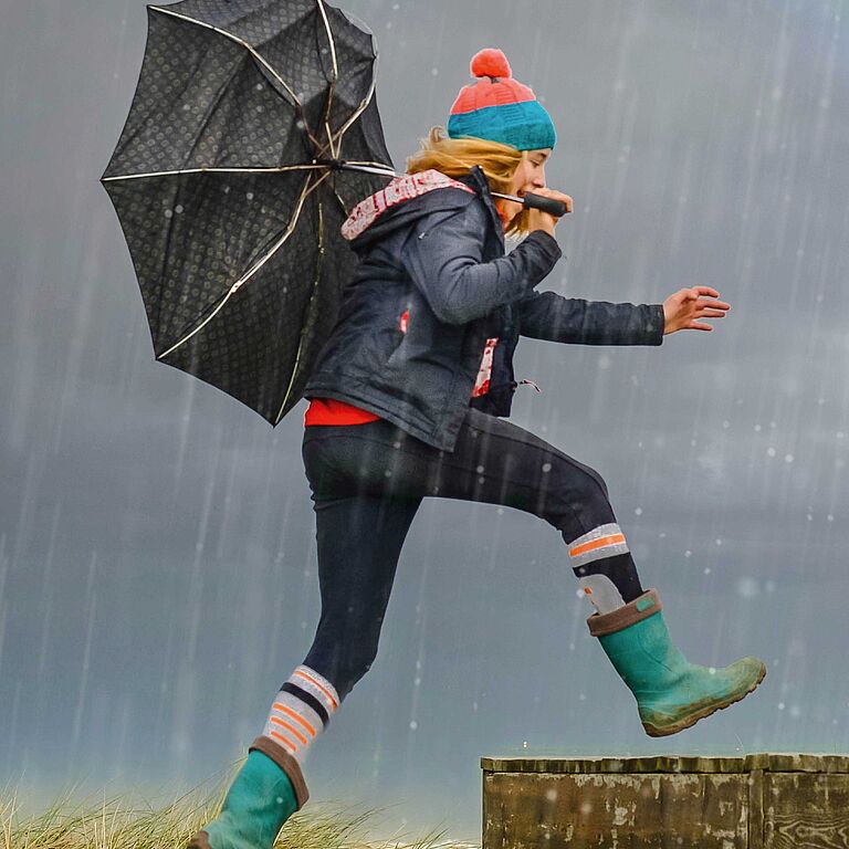 Frau mit Regenschirm geht beschwingt durch den Regen, Regenbogen am Horizont