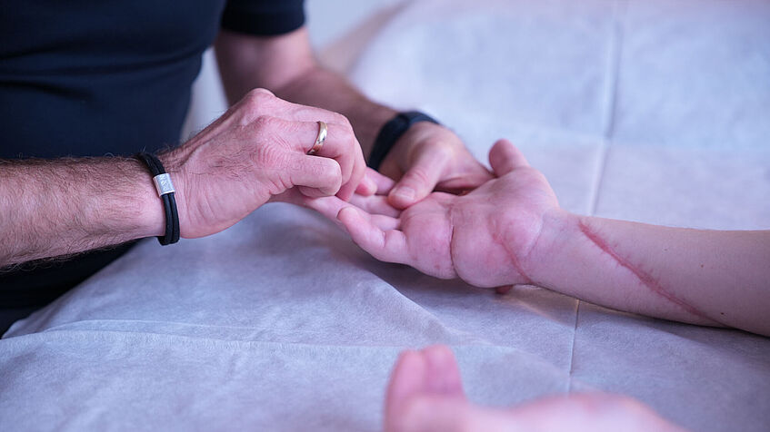 Zwei verletzte Hände liegen ausgestreckt auf einer Behandlungsbank. Zwei Hände, die rechte mit Ring und Armband, behandeln die Finger der rechten Hand. 