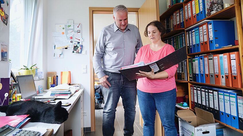 Ein Mann und eine Frau stehen in einem Arbeitszimmer vor einem Regal mit Aktenordnern und haben einen Aktenordner in der Hand.