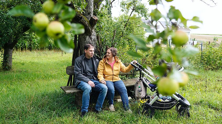 Ehepaar mit Kinderwagen sitzt auf einer Bank unter einem Apfelbaum.