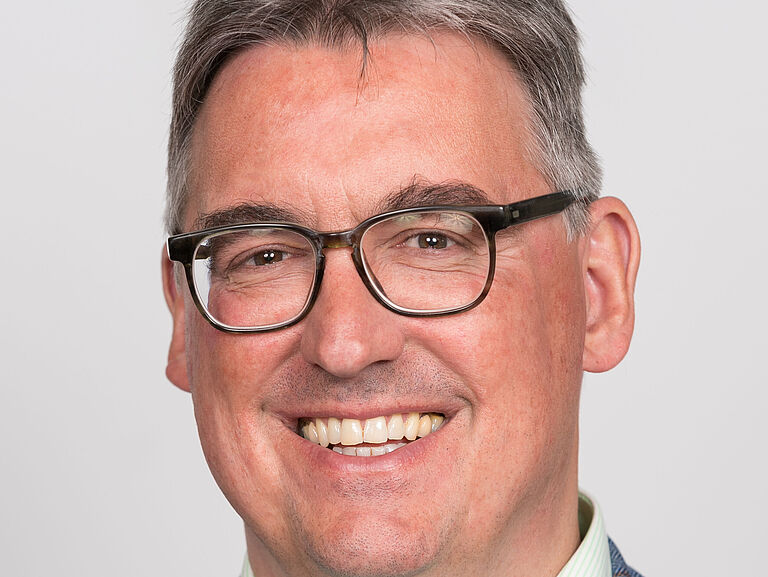 Ein Porträtfoto von Martin Kögler: Er trägt ein weißes Hemd und blaues Jackett und schaut freundlich in die Kamera. 