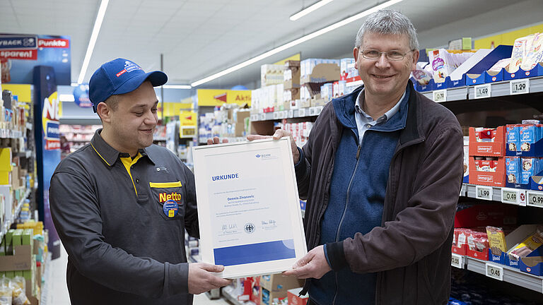 Dennis Zivanovic von Netto Marken-Discount in Arbeitskleidung erhält in einer Filiale eine Urkunde von Peter Struck überreicht. Beide lächeln zufrieden. Im Hintergrund sind Regale mit verschiedenen Produkten und Preisschildern zu sehen.