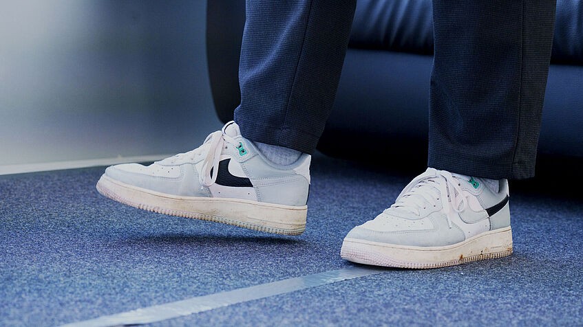 Koordinationsübungen: Ein Paar Füße in weiße Turnschuhen stehen auf blauem Teppichboden. Zwischen den Füßen ist eine weiße Linie aufgeklebt. Der rechte Fuß ist vorne angehoben. 