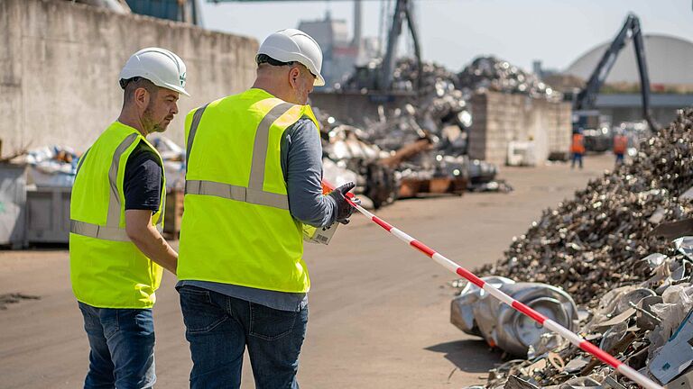 Zwei Beschäftigte mit Warnwesten und Schutzhelmen sichern auf einem Schrottplatz einen Bereich mit Absperrband ab; im Hintergrund liegen Schrottberge und Maschinen.
