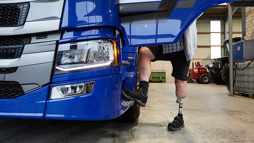 Man sieht die rechte vordere Front eines blauen Lkw. Die Tür ist geöffnet. Darunter sieht man zwei Beine im Ausschnitt - beim Einstieg in das Lkw-Fahrerhaus. Das linke Bein trägt eine Unterschenkelprothese.