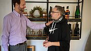 Peter Gerharz (links) und Steven Wedhorn stehen sich gegenüber. Gerharz hat seine linke Hand auf Wedhorns Schulter gelegt und lächelt ihn an. Im Hintergrund ist ein dekoratives Regal mit Weinflaschen zu sehen.