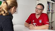 Steven Wedhorn im roten Trikot sitzt an einem Tisch und schaut seine Logopädin Wiebke Kahm an. Sie ist seitlich von hinten zu sehen. Ihre Haare hat sie zum Zopf zusammengebunden. 