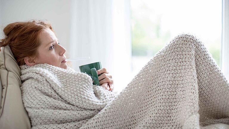 Frau liegt mit Decke auf dem Sofa, hält eine Tasse und misst Fieber mit einem Thermometer.
