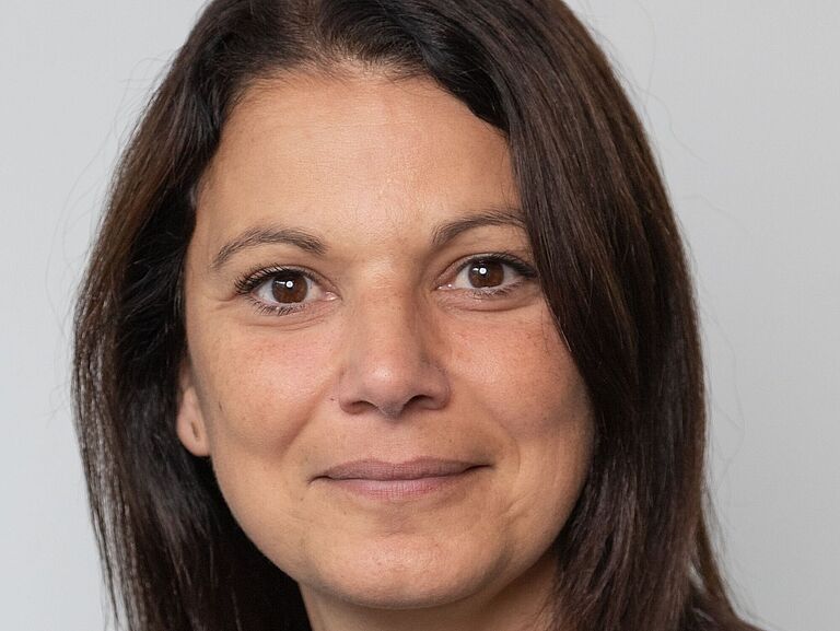 Eine Frau mit braunen langen Haaren, im Porträt fotografiert, schaut freundlich in die Kamera. 