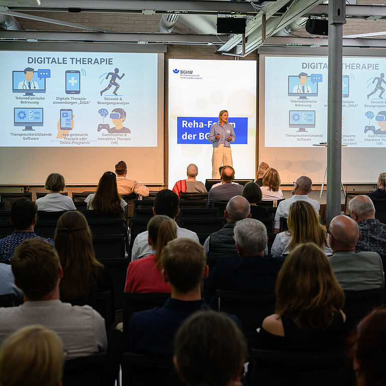 Blick ins Plenum des Reha-Forums: Eine junge Frau steht gestikulierend auf der Bühne, mit dem Gesicht zur Kamera. In der Präsentation hinter ihr kann man "Digitale Rehabilitation" lesen. Im Vordergrund sind die Rücken der Zuschauer zu sehen, die vor ihr sitzen. 